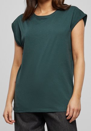 Vrouw die een donkergroene mouwloze top draagt met opgerolde schouders en donkergrijze broek tegen een effen grijze achtergrond.