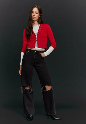 Giovane donna che indossa una giacca rossa corta con zip, top stratificati, jeans neri a vita alta strappati e tacchi neri con punta a punta, su sfondo scuro.