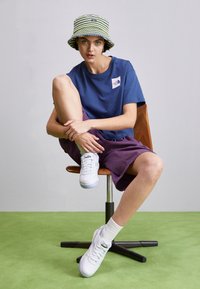 Blue t-shirt with logo, purple shorts, white sneakers with accents, and patterned bucket hat. Sitting on a wooden chair atop green flooring.