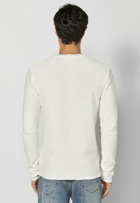 Witte, lange mouwen, gebreide shirt met een gestructureerd, ribbelpatroon. De achterkant heeft een ronde hals en aansluitende mouwen, gecombineerd met een blauwe jeans.