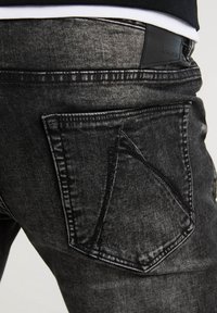 Vaqueros de mezclilla negra con un acabado desgastado, que presentan un diseño distintivo de bolsillo trasero en ángulo y una etiqueta de cuero en la cintura.