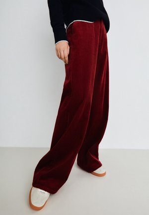 Osoba ubrana w szerokie czerwone spodnie sztruksowe, czarny sweter i białe trampki z brązowymi podeszwami, stojąca na tle jednolitego jasnego tła.