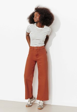Femme aux cheveux afro portant un t-shirt blanc à rayures, un pantalon large couleur rouille et des baskets à imprimé léopard, debout contre un mur blanc.