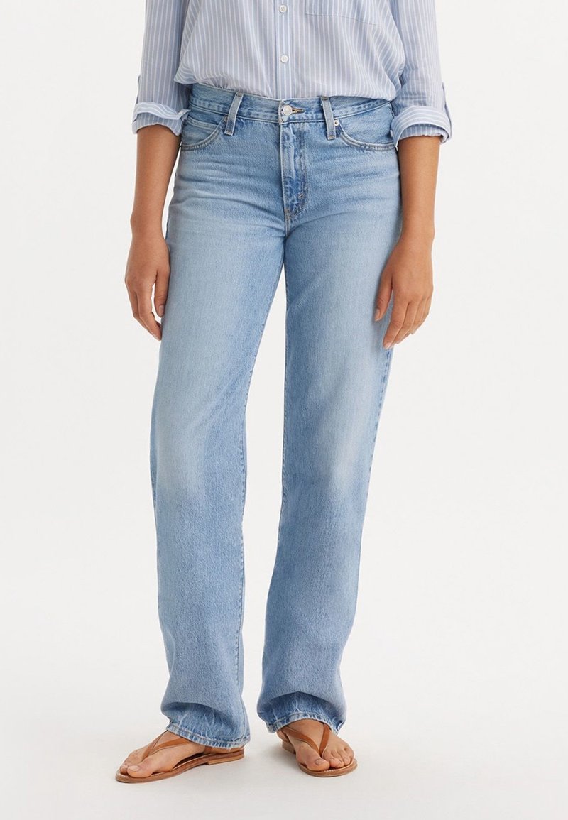 Femme portant un jean bleu clair à jambes droites, une chemise rayée rentrée avec les manches retroussées, et des sandales tongs marron, debout sur un sol blanc.