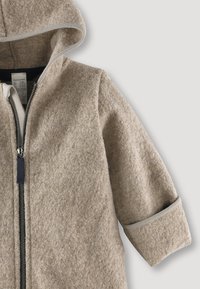Sudadera beige con cremallera hecha de una tela suave y texturizada, con capucha, puños enrollados y cremallera oscura en contraste. Diseño simple y funcional.