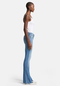 Jeans en denim bleu évasés avec un délavage clair, présentant une taille mi-haute, une coupe ajustée et des coutures contrastantes. Portés avec un débardeur blanc.