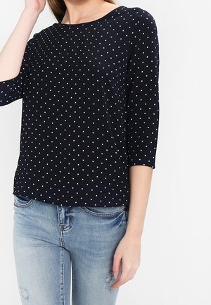 Femme portant un haut bleu marine à manches trois-quarts avec des pois blancs et un jean bleu clair déchiré sur fond blanc.