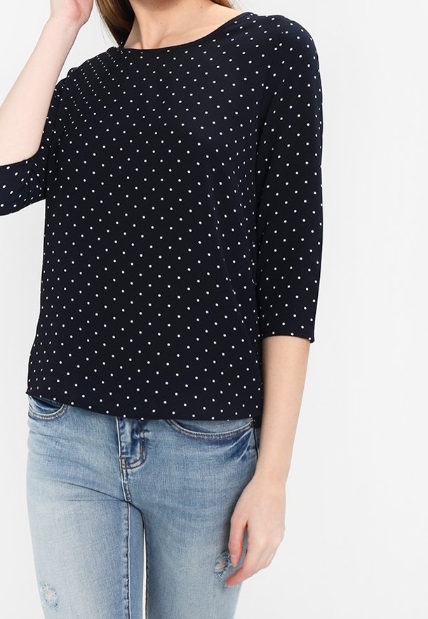 Femme portant un haut bleu marine à manches trois-quarts avec des pois blancs et un jean bleu clair déchiré sur fond blanc.