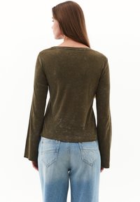 Top de manga larga verde oliva con cuello barco, que presenta una tela suave y texturizada. Combinado con jeans de tiro alto azul claro, que muestran un diseño sencillo en la parte posterior.