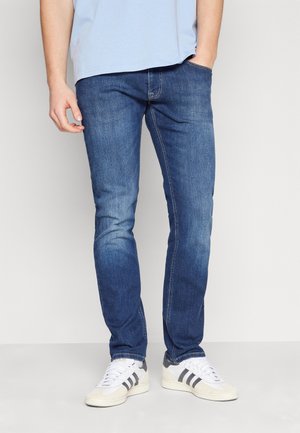 Man draagt blauwe slim-fit jeans met vervaagde details en witte sneakers met zwarte strepen, één hand in de zak, staand tegen een effen achtergrond.