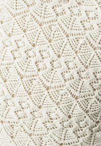 Pletená textilie v slonovinové barvě s diamantovým vzorem. Krajkovitý design má složité detaily a měkký, texturovaný vzhled.