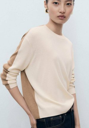 Young woman wearing beige and brown color-block sweater and dark jeans, with gold square hoop earrings, standing against a white background.