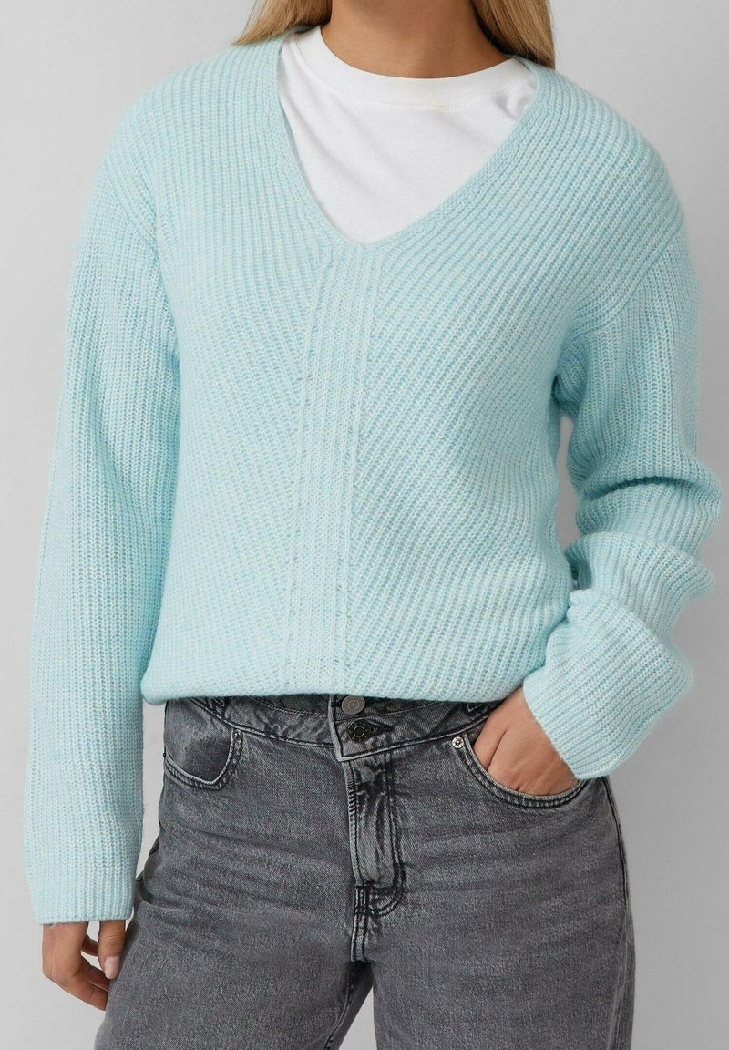 Femme portant un pull en maille bleu clair par-dessus une chemise blanche et un jean noir délavé taille haute, une main dans la poche.
