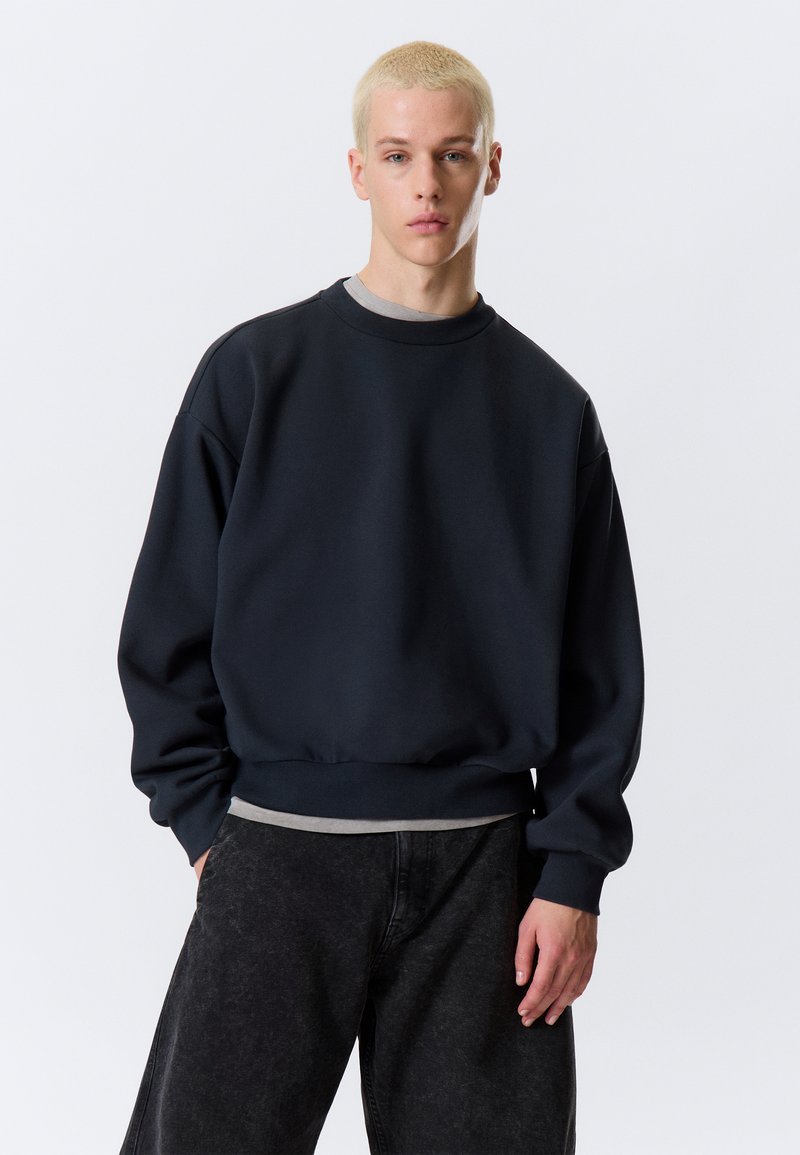 Dark navy sweatshirt with a crew neckline, ribbed cuffs, and hem. Worn over a light gray shirt, paired with dark gray pants.