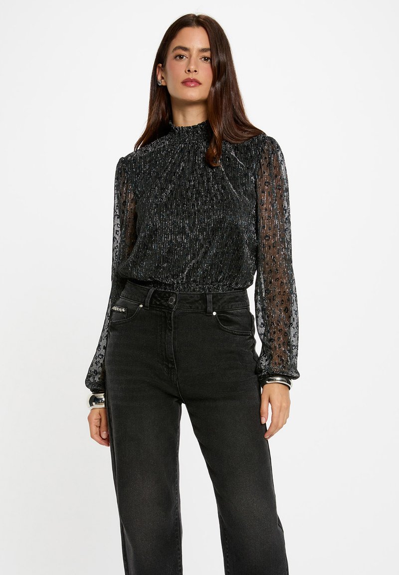 Zwarte langemouwtop met een hoge hals, voorzien van een gestructureerde glans en doorzichtige mouwen. Gedirigeerd met hoge taille zwarte jeans.