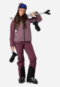 Skidragt bestående af en lilla softshell-jakke, matchende bukser med sorte detaljer og sorte skistøvler. Tilbehøret inkluderer en beanie og skibriller.