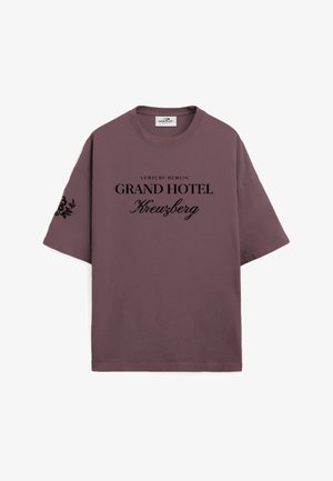 Młody mężczyzna z krótkimi włosami, ubrany w luźny fioletowy T-shirt z czarnym napisem i białe spodnie, stoi z rękami w kieszeniach na jednolitym tle.