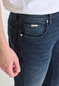 Jeans de mezclilla azul oscuro con un bolsillo frontal, que presentan una pequeña etiqueta metálica como acento. La tela tiene una textura suave y un ligero desvanecimiento.