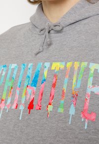 Gros plan d'un sweat à capuche gris avec des lettres multicolores dégoulinantes formant le mot « FORTNITE » sur la poitrine, porté par une personne à la peau claire et aux cheveux bouclés.
