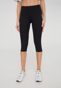 Leggings neri fino al ginocchio, realizzati in materiale elastico, presentano una texture liscia e un minimo dettaglio di cuciture. Indossati con scarpe da ginnastica bianche.