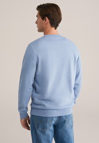 Vīrietis ar brūniem matiem, ģērbies gaiši zilā sporta kreklā un zilos džinsos, stāv pretēji neitrālai bēšai fona.