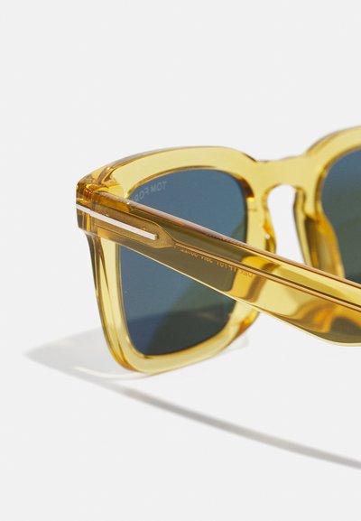 Kollased läbipaistvad prillid ruudukujulise raami, tumeda varjundiga läätsed ja kuldsed aksessuaarid templites. Sile pinna tekstuur.