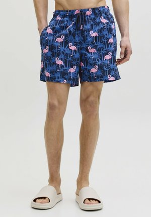 Homme portant un short de bain bleu marine avec un motif de flamants roses et de palmiers, sandales beige à enfiler, debout les mains détendues.