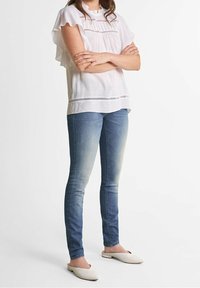 Blouse blanche avec des manches volantes et des détails en dentelle, assortie à un jean ajusté bleu clair et des chaussures slip-on beiges.