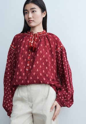 Junge Frau trägt eine lockere rote gemusterte Bluse mit Quasten und hellbeige Hose, steht vor einem schlichten hellen Hintergrund.