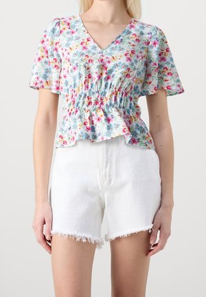 Vrouw draagt een bloemen blouse met korte mouwen en V-hals, met een geplooide taille en witte versleten denim shorts tegen een effen achtergrond.