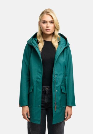 Vrouw met blond haar die een blauwgroene capuchonjas draagt over een zwart shirt en donkere spijkerbroek, staand tegen een effen witte achtergrond.