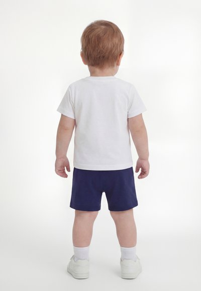 Νήπιο με ανοιχτό καστανά μαλλιά στέκεται γυρισμένο προς τα πίσω, φορώντας λευκό μπλουζάκι, σκούρο μπλε σορτς, λευκές κάλτσες και λευκά αθλητικά παπούτσια σε λευκό φόντο.
