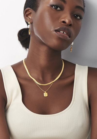 Joias de ouro incluem um colar grosso e ondulado e uma corrente em camadas com um pendente quadrado. Os brincos apresentam um design de gota com uma pérola.