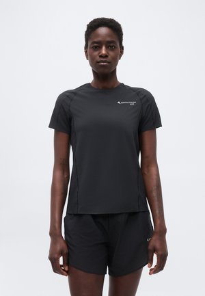 Modèle féminin portant un t-shirt de sport noir à manches courtes et un short assorti, debout devant un fond gris clair uni.