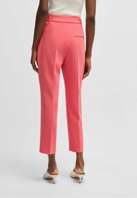 Růžové kalhoty na míru s hladkou texturou, zúženými nohavicemi a detailem kapsy na zadní straně, doplněné béžovými lodičkami na vysokém podpatku.