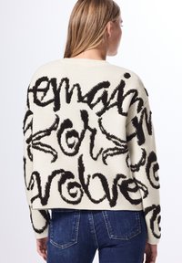 Maglione crema con lettere nere testurizzate, vestibilità comoda, spalle basse e polsini a costine, abbinato a jeans in denim blu scuro.