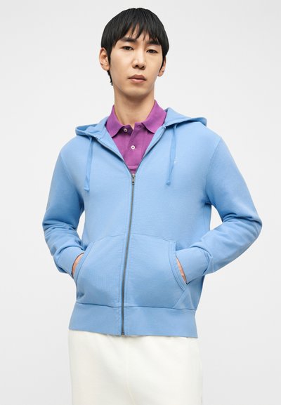 Άνδρας που φορεί ανοιχτό μπλε φούτερ με φερμουάρ πάνω από μωβ πουκάμισο με γιακά και λευκό παντελόνι, με τα χέρια στις τσέπες, μπροστά σε λευκό φόντο.