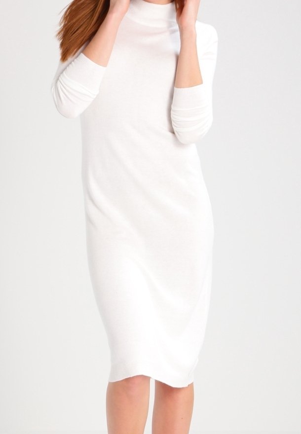 Femme portant une robe midi blanche ajustée à manches longues et col montant, debout devant un fond clair uni.