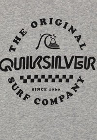 Šedá bavlněná tkanina s černým kruhovým logem, text zahrnuje "Quiksilver" a "The Original Surf Company," s kostkovanými akcenty.