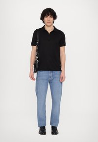 Μαύρο πόλο κοντομάνικο με κολάρο που κλείνει με φερμουάρ, συνδυασμένο με ανοιχτό μπλε τζιν και μαύρα παπούτσια. Το μοντέλο κρατά μια μαύρη τσάντα ώμου.