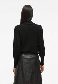Femme aux longs cheveux noirs portant un pull noir à manches bouffantes et une jupe en cuir noir, se tenant de dos devant un fond blanc uni.