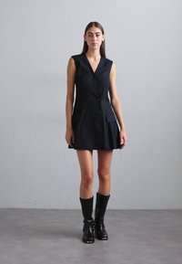 Rochie fără mâneci de culoare albastru marin, cu un design cu două rânduri de nasturi, două buzunare frontale, formă structurată și un material texturat. Asortată cu cizme până la genunchi.
