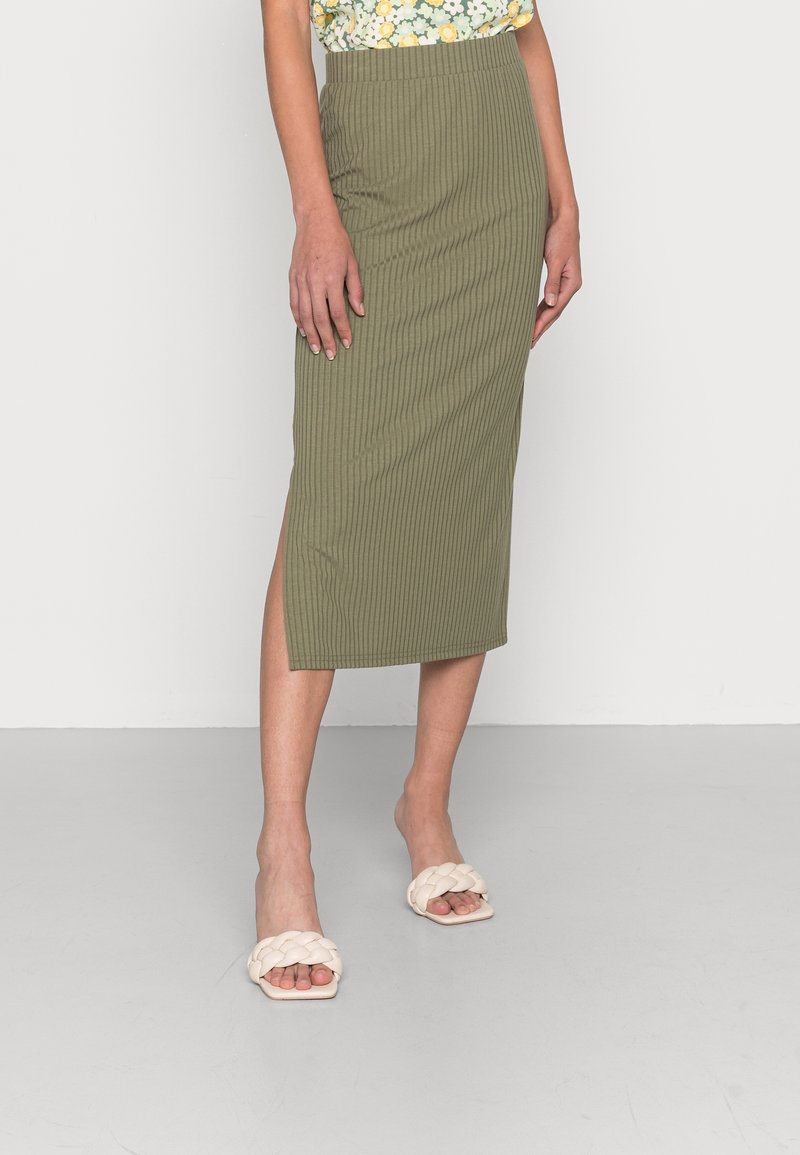 Femme portant une jupe midi côtelée vert olive avec une fente latérale, un haut fleuri et des sandales blanches tressées à bout ouvert, debout sur un sol gris.