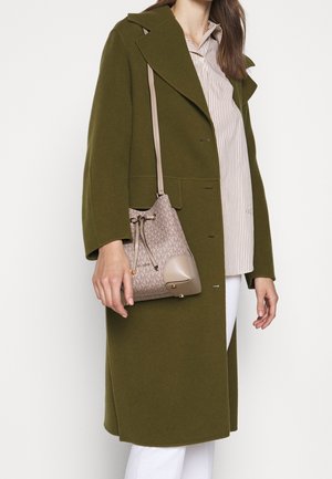 Femme portant un manteau vert olive, un pantalon blanc, une chemise rayée et portant un sac seau beige de créateur sur son épaule.