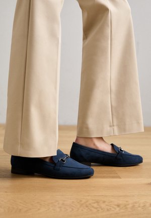 Personne portant un pantalon beige à jambes larges et des mocassins bleu marine en daim avec un détail argenté sur la boucle, debout sur un sol en bois.