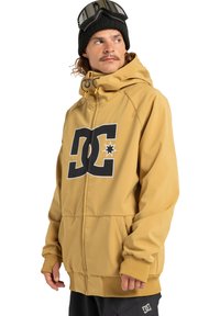 Man in mosterdgele DC-hoodie, zwarte beanie met skibril erop, en zwarte broek met DC-logo, kijkend naar de zijkant.