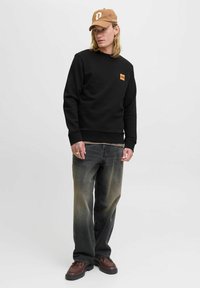 Joven vestido con una gorra color beige, sudadera negra con un pequeño logo naranja, jeans negros desgastados y zapatos marrones, de pie frente a un fondo liso.