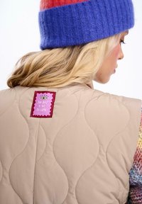 Beżowa pikowana kamizelka z falistym przeszyciem, z kolorową metką z tłem w kolorze różowym i fioletowym napisem "NAJLEPSZE JESZCZE PRZED NAMI."