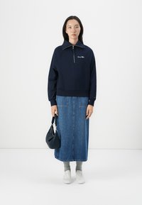 Námornícky modrá mikina na zips s vysokým golierom; v kombinácii s dlhou džínsovou sukňou. Biele tenisky a malá čierna kabelka dopĺňajú outfit.