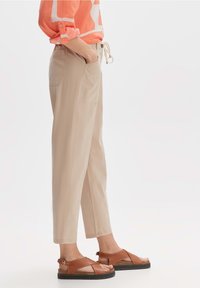 Beige Hosen mit lockerer Passform, ausgestattet mit Seitentaschen und einem Kordelzugbund, kombiniert mit braunen Sandalen im Schnürdesign.